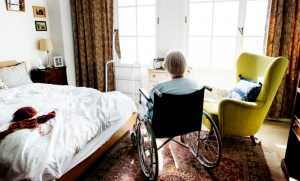 Elderly woman sitting in a wheelchair in a bedroom.