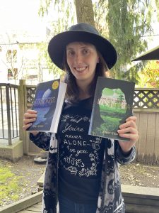 Brown-haired woman wearing a black wide-brimmed hat. She is holding two books, Dysnomia: Outcast on a Distant Moon and Dysnomia: Home Lies in Your Heart.