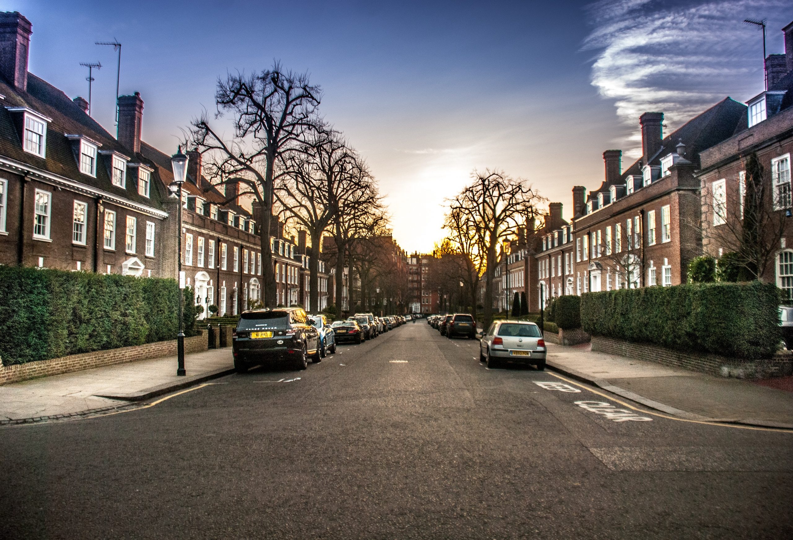 Two rows of brown houses facing each other, with cars lined up on each side of the road. The sun is setting and the trees on either side of the road are barren.