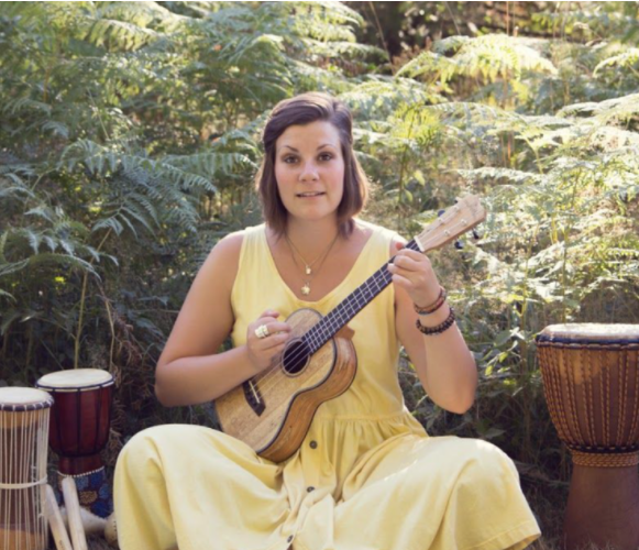 Woman in yellow dress holds guitar while sitting outdoors, with traditional drums on either side of her