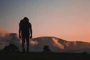 A person with a backpack, facing away from the camera, hiking up a hill during sunset.