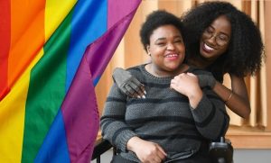 Close-up of two disabled Black people smiling and holding hands. The woman in the back is wearing compression gloves and looking lovingly at the non-binary person in the front, who’s sitting in a power wheelchair. There is a graphic of a rainbow pride flag covering the left side of the image.