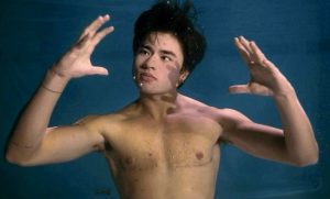 Actor Chella Man is underwater in a pool, arms out, with his shirt off. Bubbles float past his face