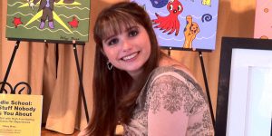 A brown-haired woman with bangs smiles at the camera, with her cartoon art behind her