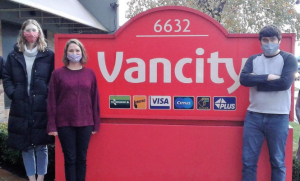 Two women and a young man, Justin Ferrano, stand outside next to a large Vancity sign. They are all wearing face masks.