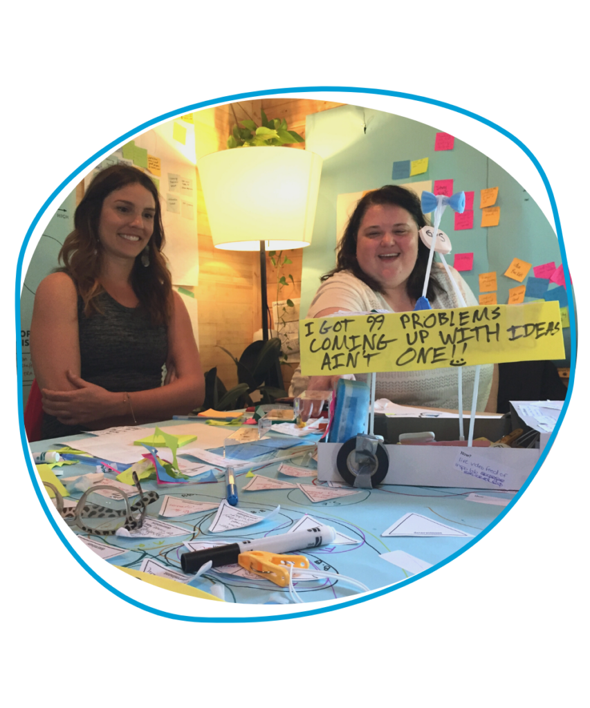 Two women at a table covered with colourful sticky notes and papers. There is a small sign on the table which reads "I got 99 problems. Coming up with ideas ain't one!"