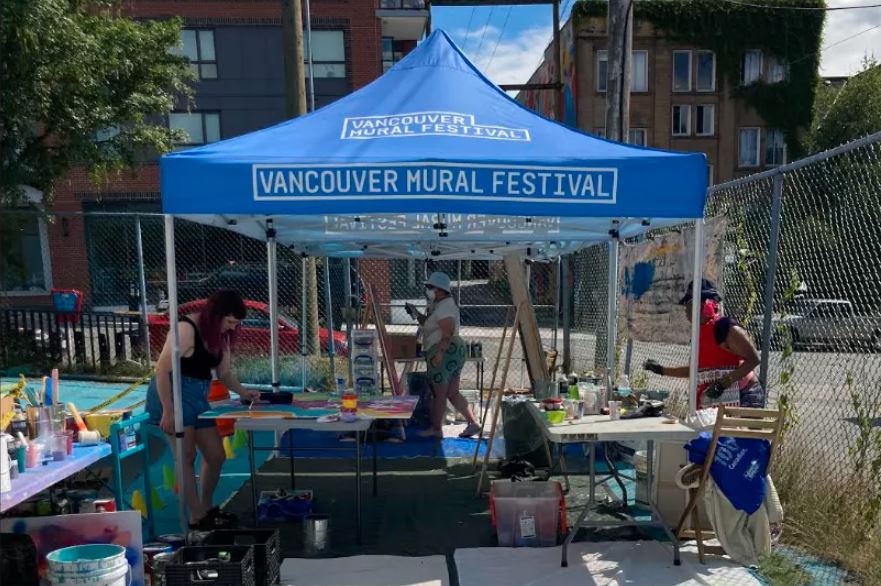 Artists working outside under a blue Vancouver Mural Festival tent