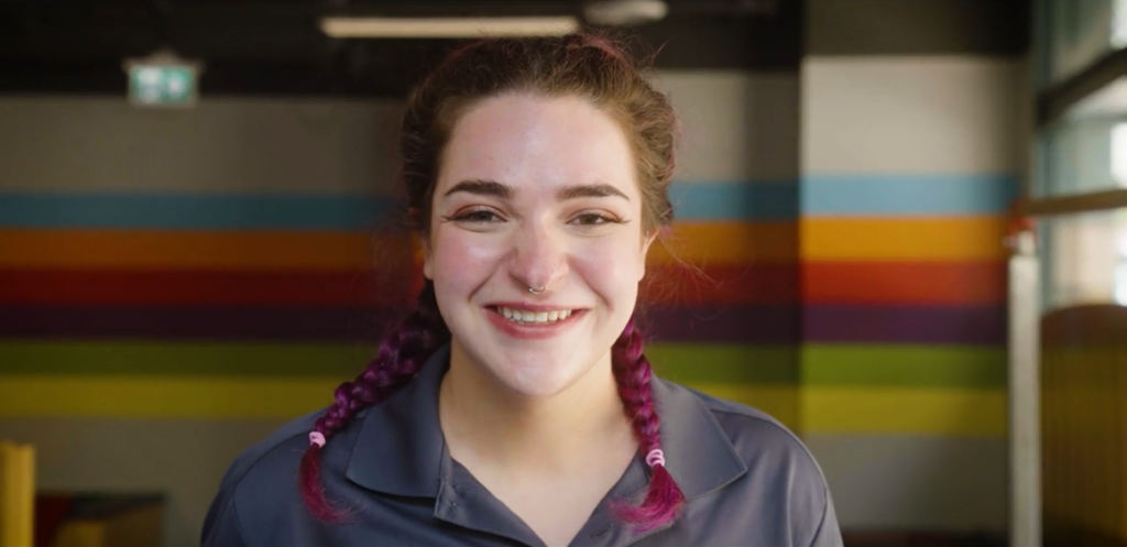 A smiling young white woman with her hair in pigtails. She is standing in a large room with colourful horizonal stripes painted on the wall behind her.