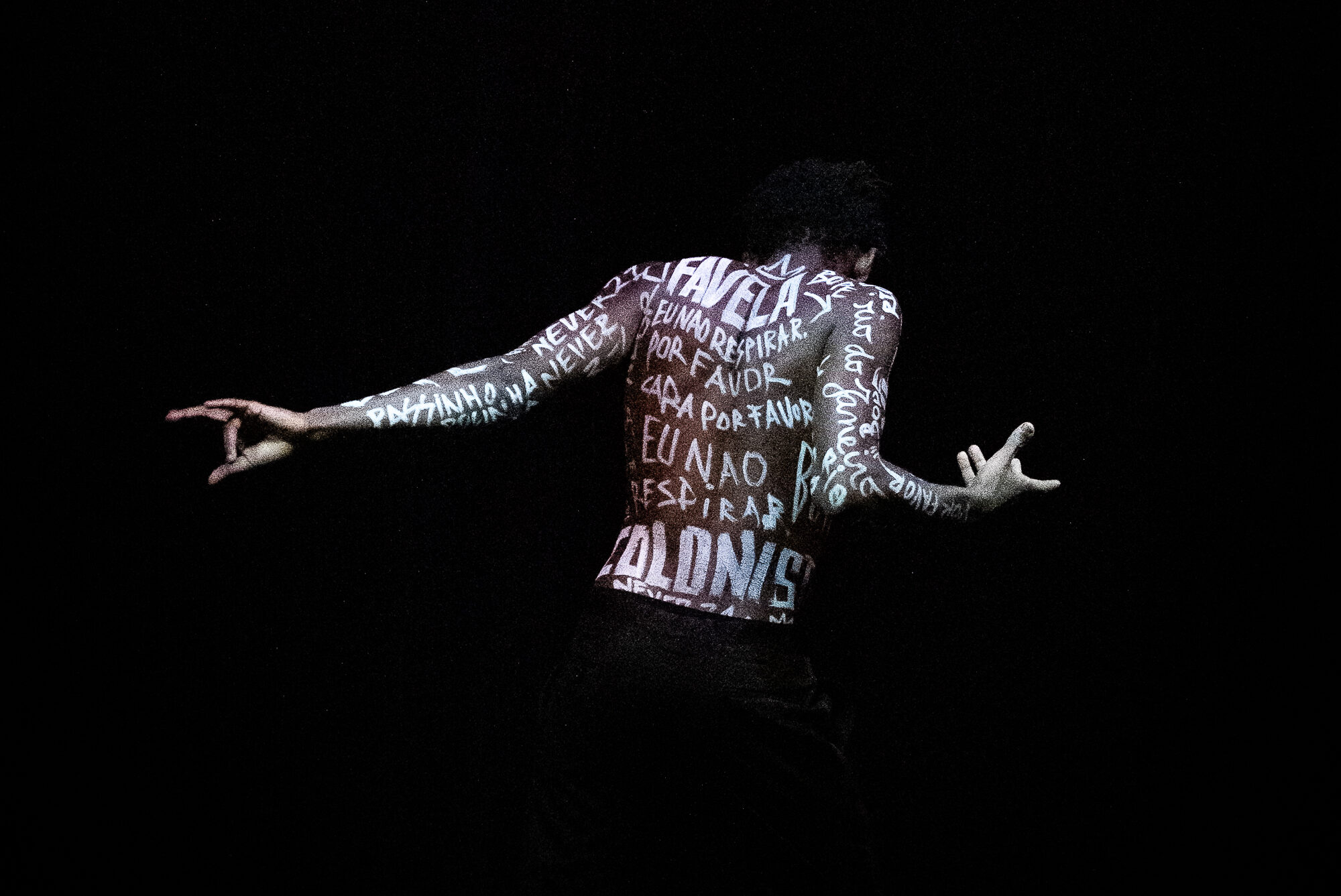 A dancer in black pants stands facing away from the camera, arms gesturing. The room is darkly lit, highlighting their bare back and arms which are covered in white-painted testimonials of the families of young Black men who have been victims of gun violence.