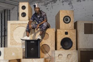 nora chipaumire, wearing a white and black-patterned jumpsuit, thick black necklaces, white and black head covering, and white sneakers, looks into the camera. she sits on top of a high stack of wood stereo speakers, feet resting on top of a black speaker.